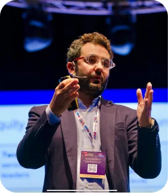 Antonio Bebba in a plum suit and conference lanyard, both hands open mid-sentence on a blue-lit stage.