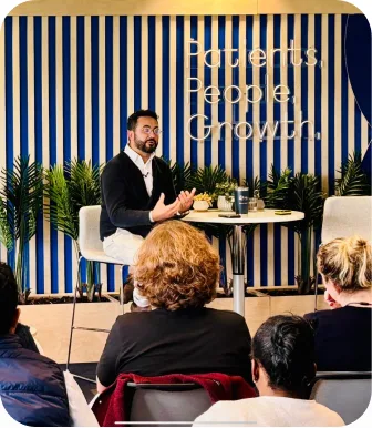 Antonio Bebba in conversation on a low stage in front of a slatted blue backdrop, the front rows of the audience in shot.