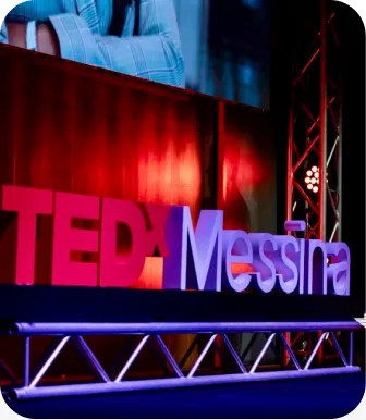 The TEDxMessina sign lit pink and blue on a stage hung with red curtains.