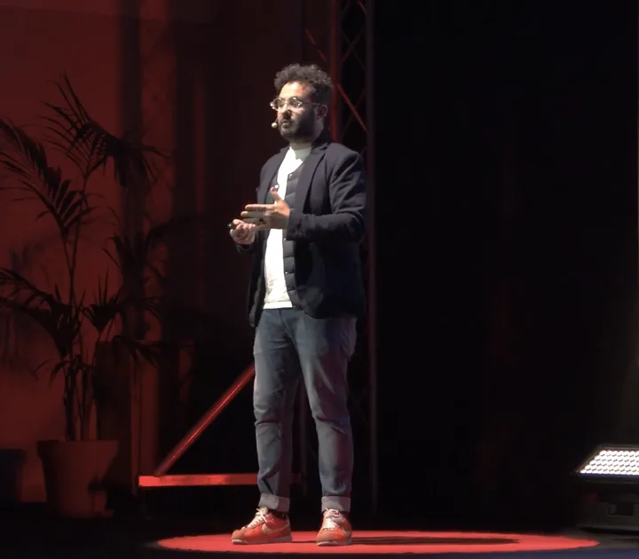 Antonio Bebba mid-sentence on the TEDxMessina stage, lit red, a palm behind him.